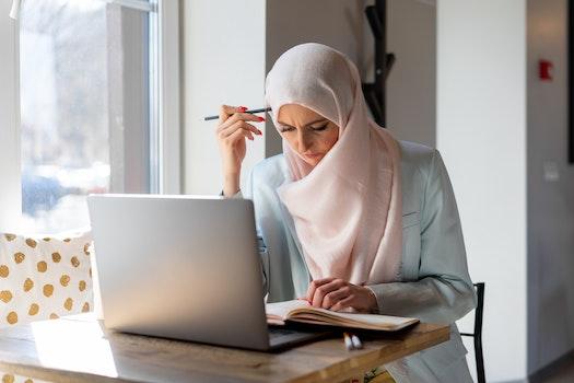 A teacher prep candidate sits at her laptop studying for her Texas certification exams. She is wearing a hijab and holding a pencil against her head while she reads.